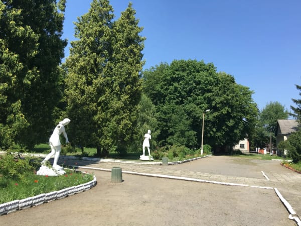 Дві білі скульптури людей у Печерському парку, оточені зеленою травою, квітами та високими деревами під блакитним небом