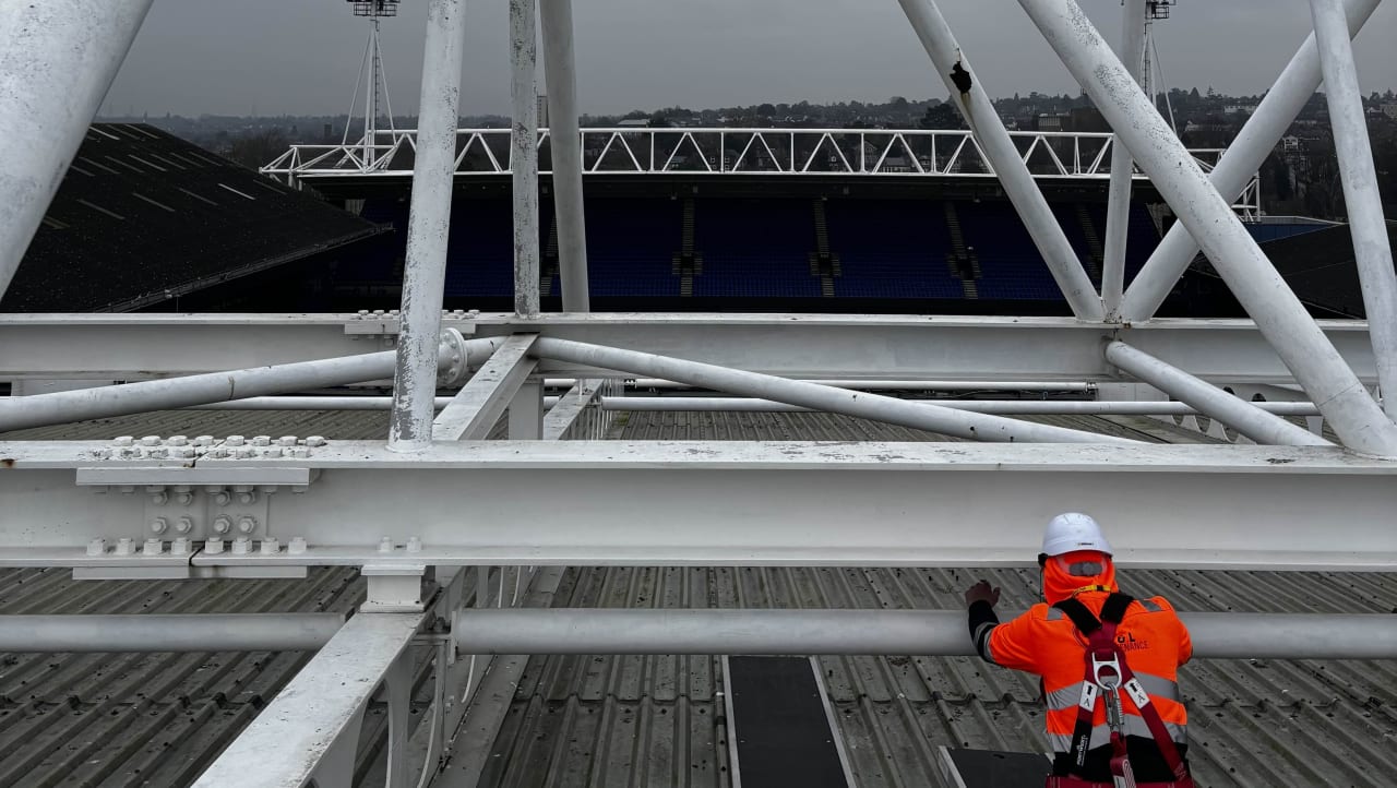 Operative in a harness working on white stand roof steelwork above a football pitch.