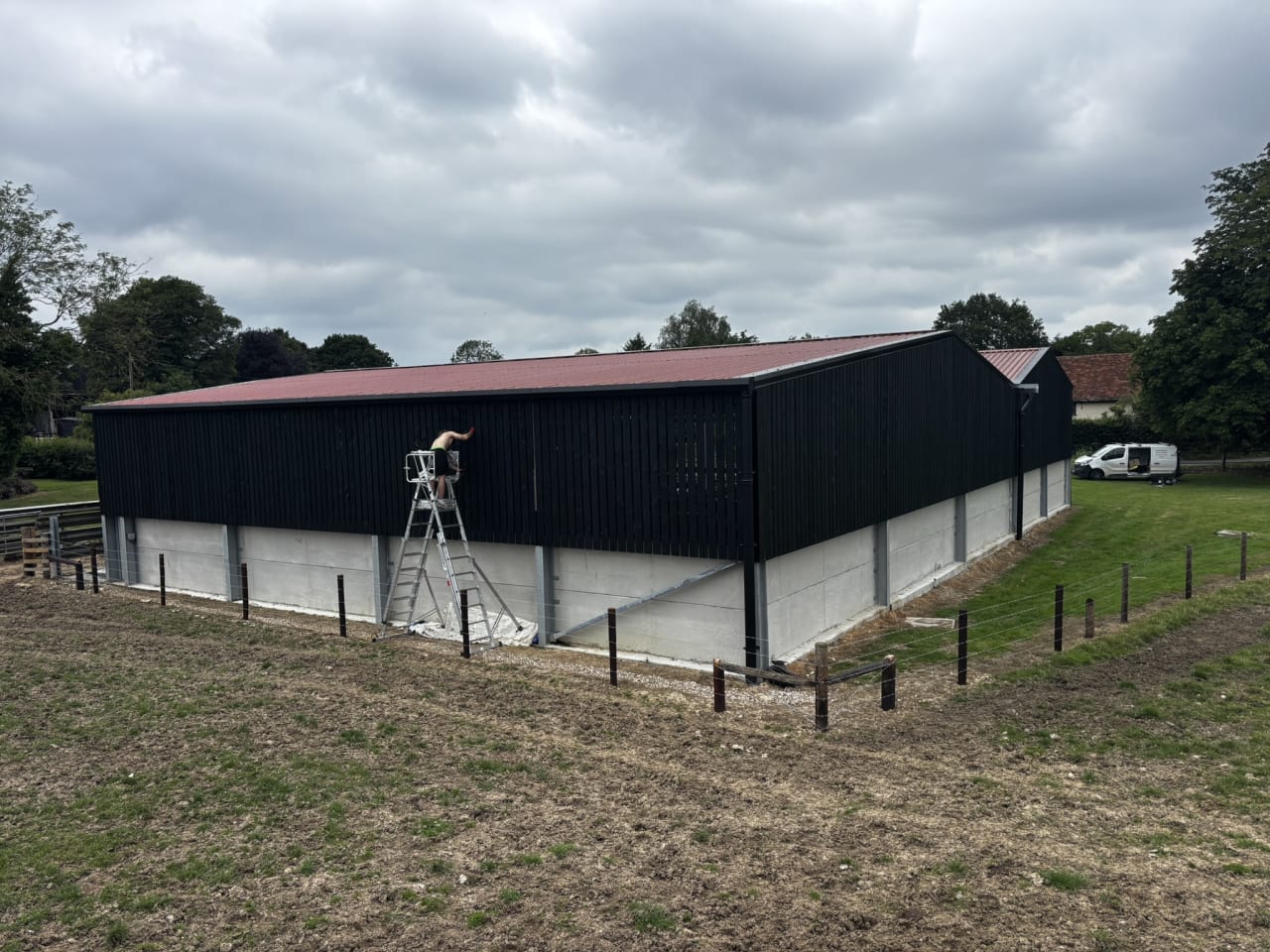 Black timber clad agricultural barn with a red roof and a tower scaffold against one wall.
