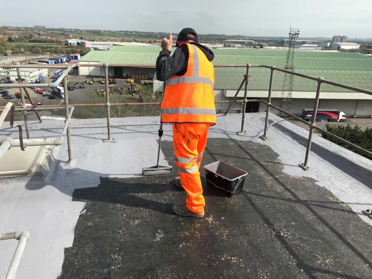Operative in orange overalls applying a grey roof coating on a flat industrial roof, with a harness line and edge protection in place.