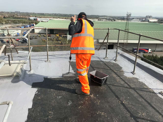 Operative in orange overalls applying a grey roof coating on a flat industrial roof, with a harness line and edge protection in place.