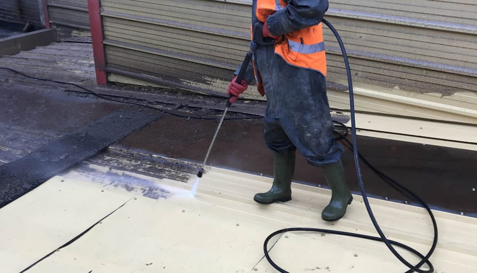 Operative in wet weather gear pressure washing a roof sheet beside profiled cladding.