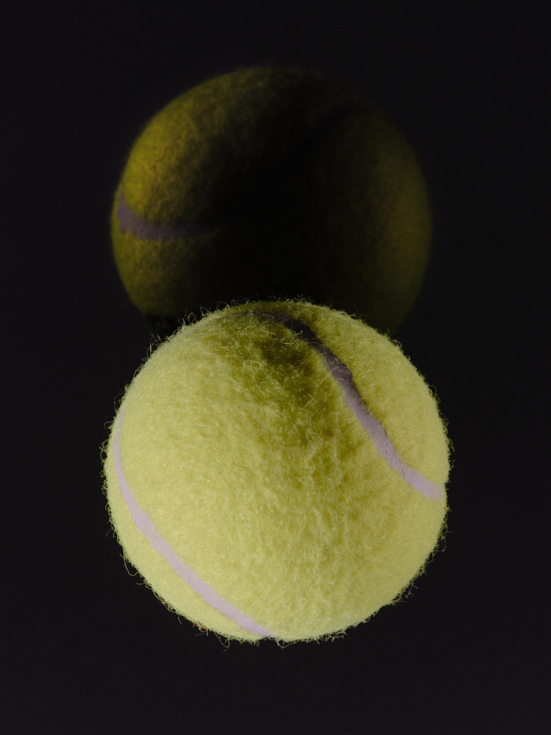 Two tennis balls in soft focus, one in shadow and one lit sharp yellow-green, against a pale gray background.