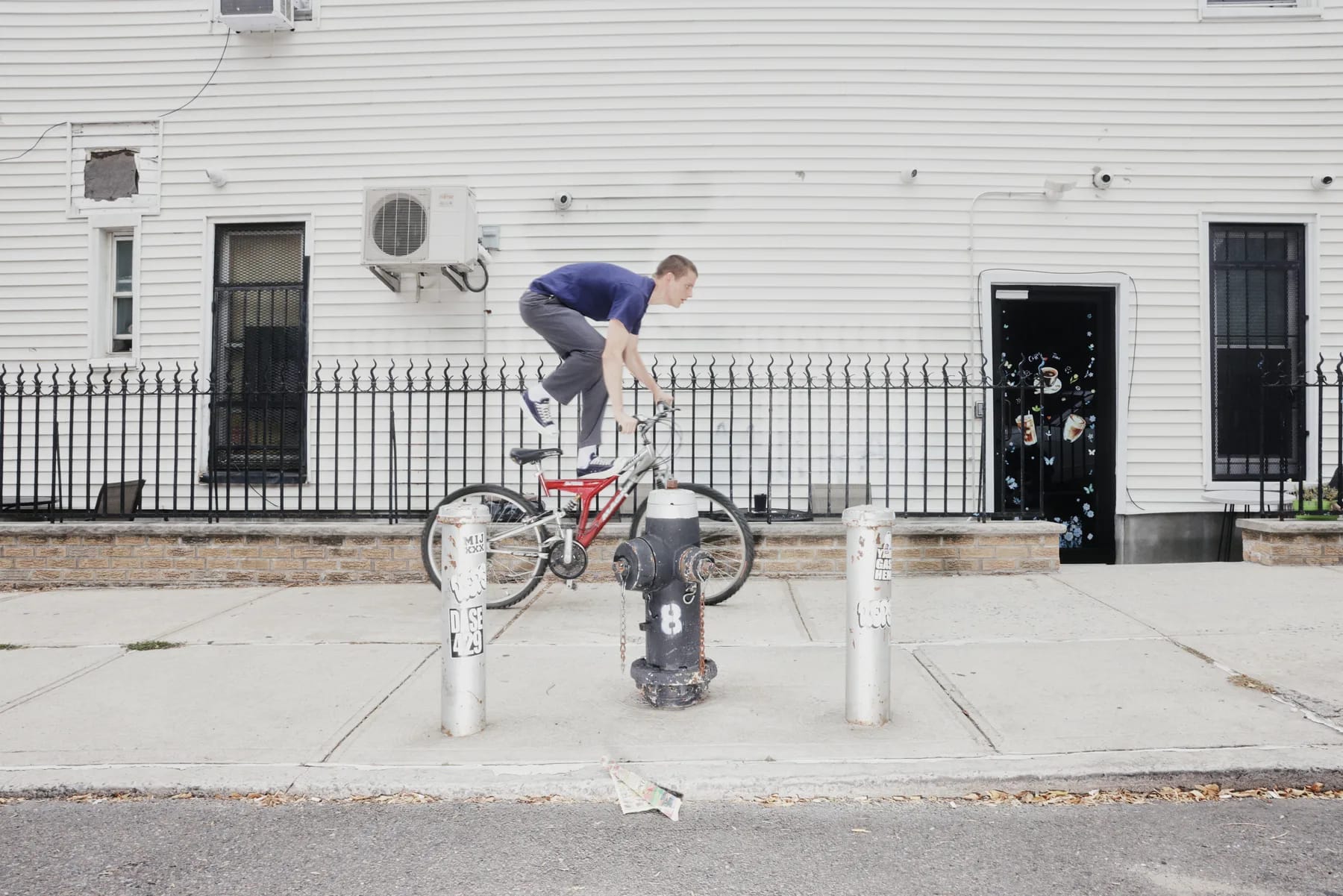 A cyclist hopping a red BMX bike over a fire hydrant outside a white clapboard row house.