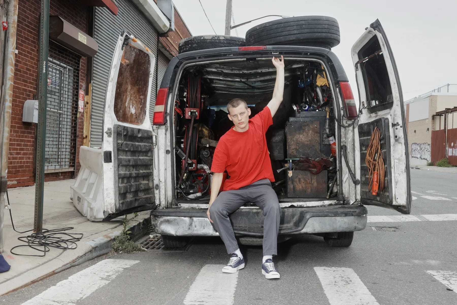 A figure in a red tee sitting in the open back doors of a battered work van loaded with tires.
