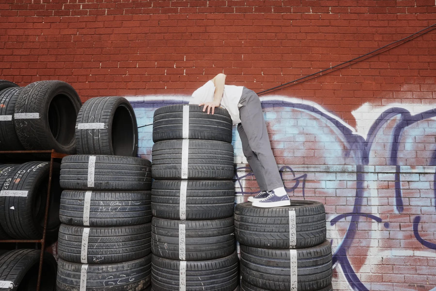 The same figure draped face down over a stack of tires against a graffitied red brick wall.