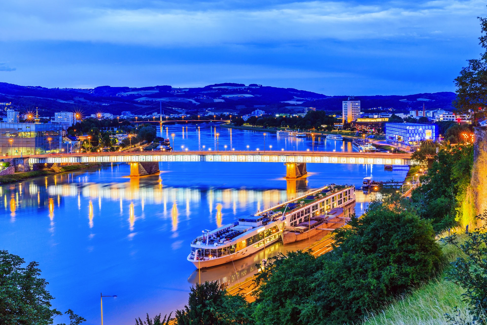 Flusskreuzfahrtschiffe an einer beleuchteten Brücke in einer Stadt bei Dämmerung.