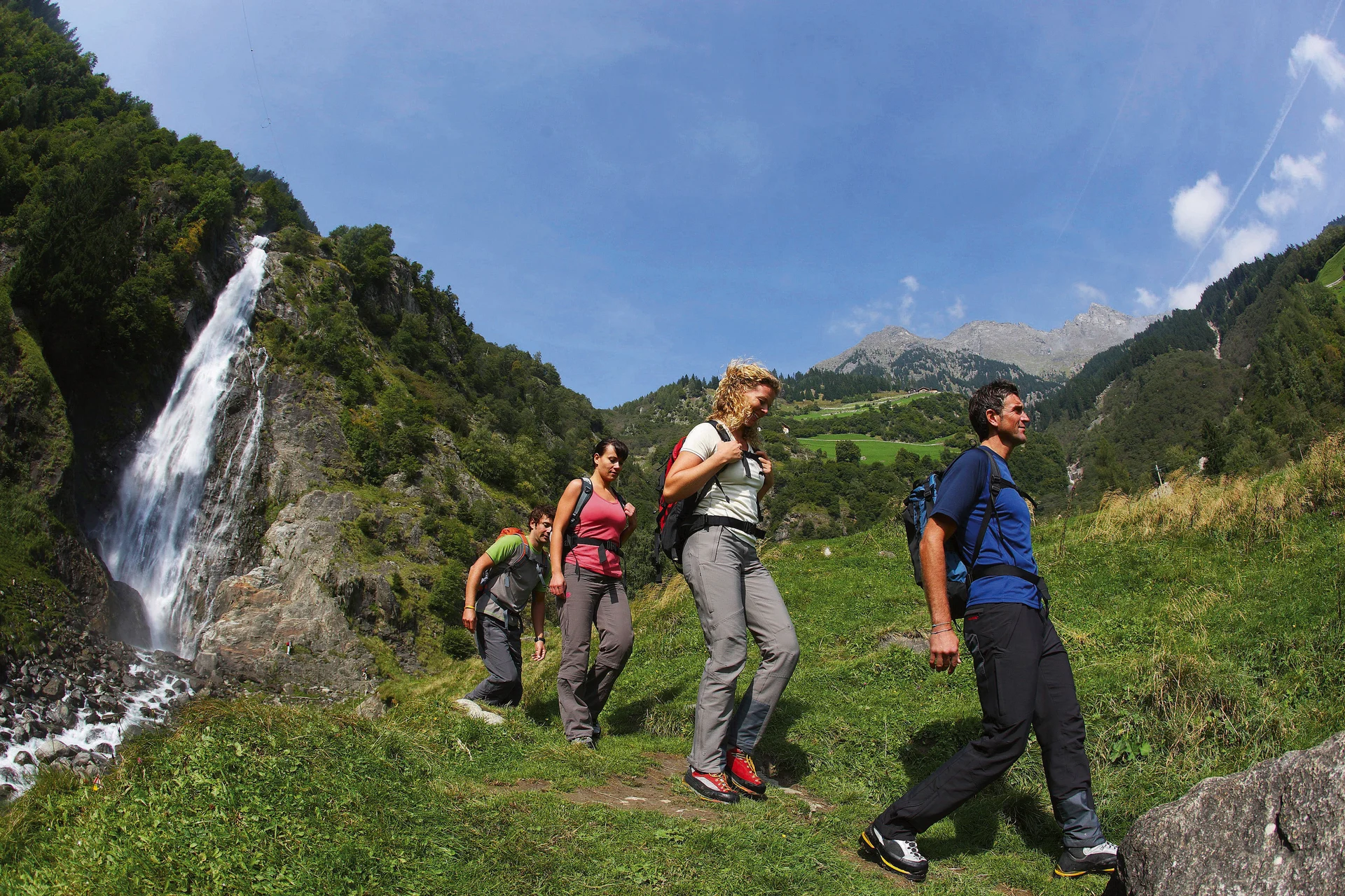 Gruppe von Wanderern auf einem Pfad im Gebirge mit Wasserfall im Hintergrund.