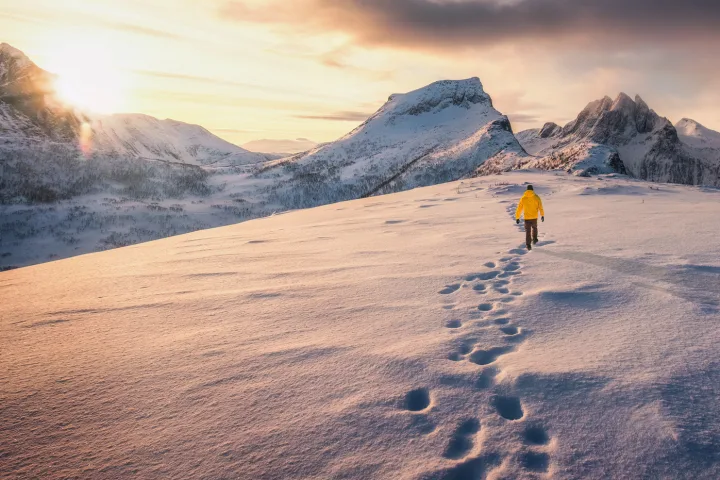Person wandert über verschneite Landschaft mit Bergen im Hintergrund bei Sonnenuntergang.