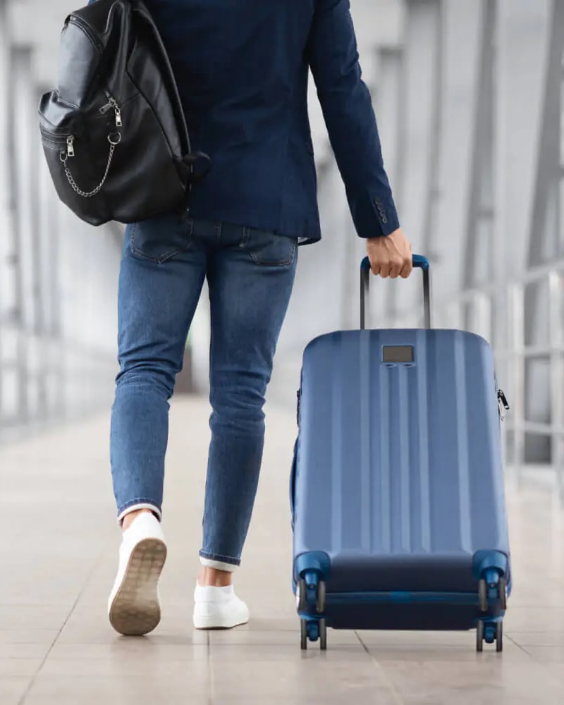 Person von hinten zieht einen blauen Rollkoffer durch einen Flughafengang