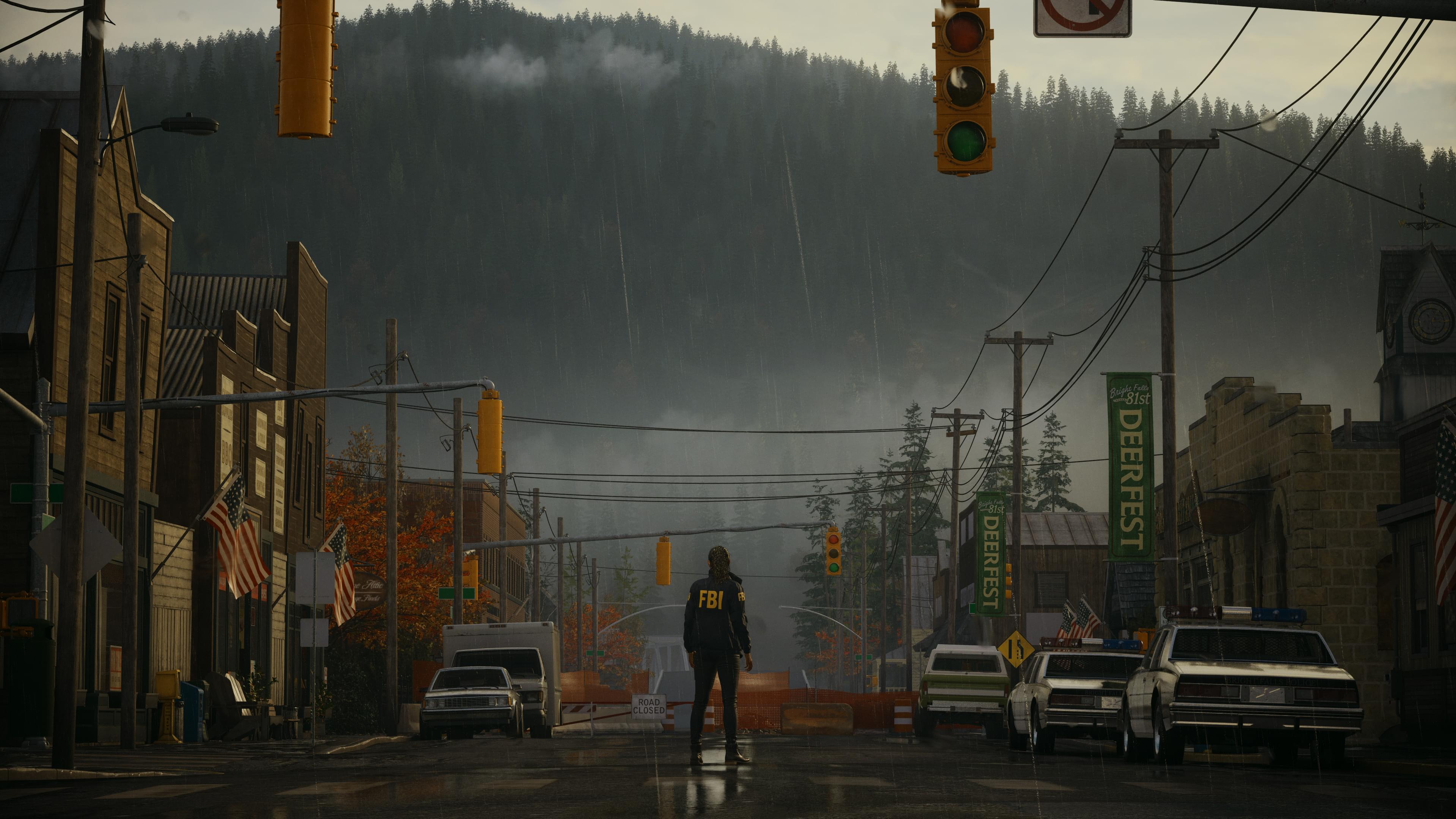 A screenshot of the video game Alan Wake II showing Bright Falls. There is a mountain in the backdrop with a foggy overcast. There is a woman standing in the middle of a street. The left side is lined with small, historic looking buildings. The right side is lined with cars.