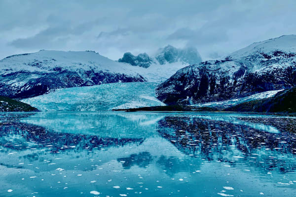 A glacier flows between snow-covered peaks, mirrored in still Patagonian water