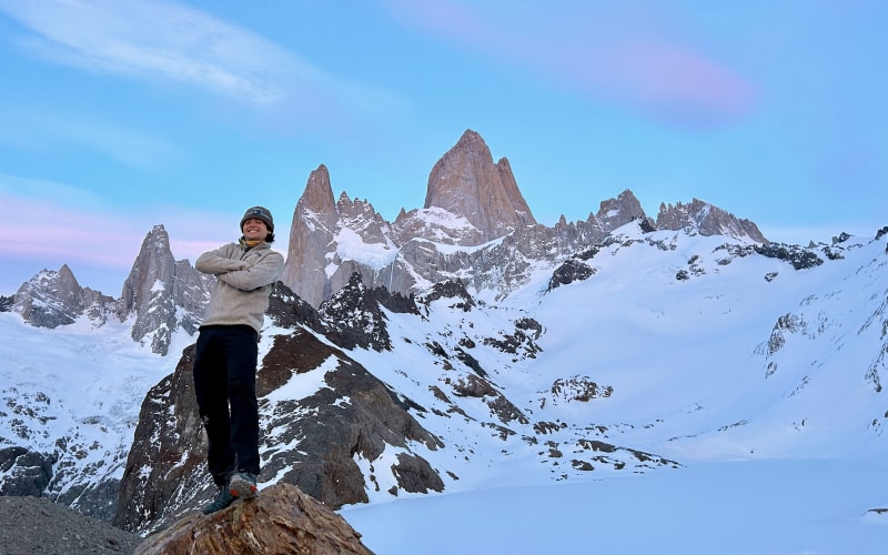 A solo traveler stands on rocky Patagonian terrain before snow-capped peaks under a clear blue sky