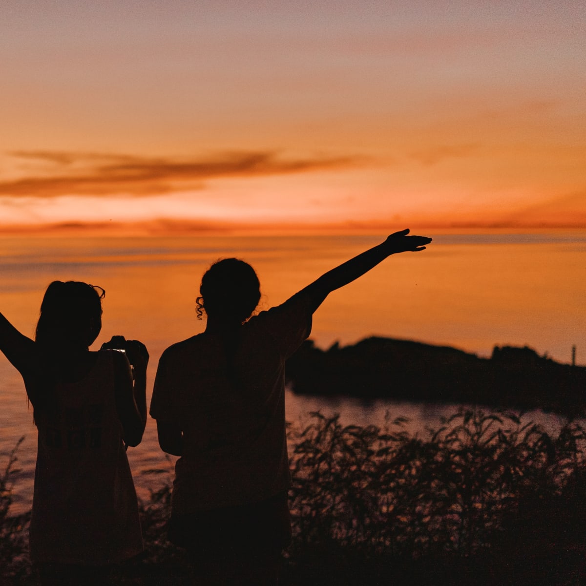 Two silhouetted figures raise their arms toward a glowing orange sunset over the sea, tall grasses lining the shore below