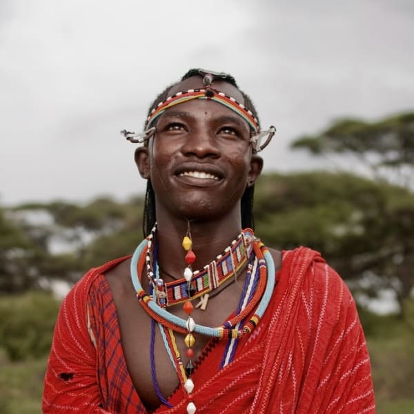 A smiling Maasai man in red dress and beadwork holds a staff on the savanna