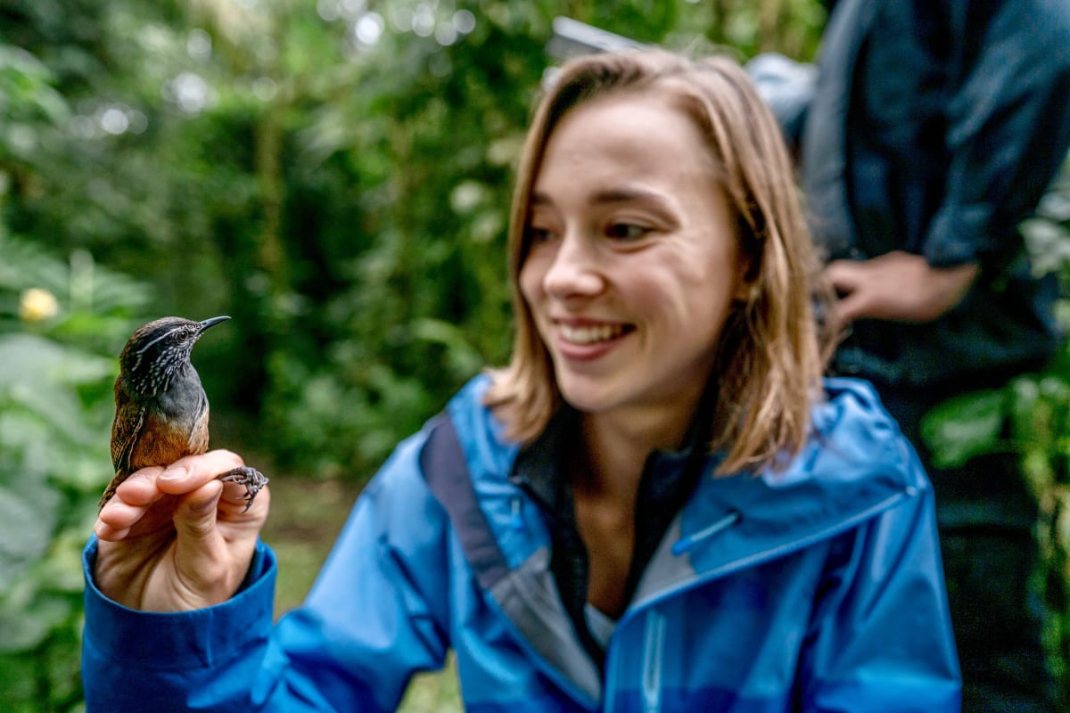 A smiling student in a blue rain jacket holds a small brown bird with a long, curved bill on her fingertips, with green forest behind her.