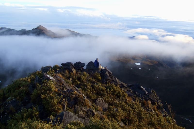 A hiker sits on a rocky summit above a sea of clouds, small lakes dotting the valley below