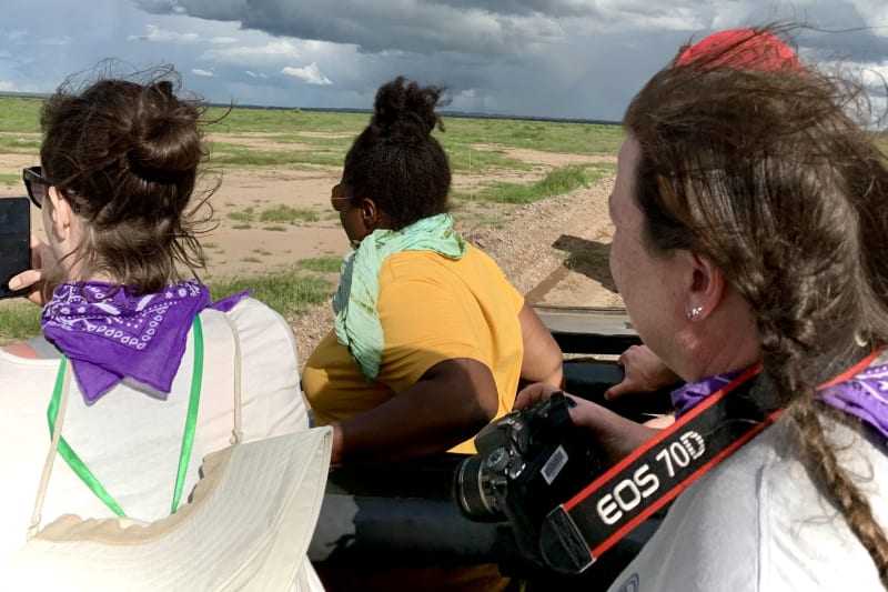 Students with cameras gather atop an open-air safari vehicle beneath storm clouds and a rainbow on the Kenyan savanna