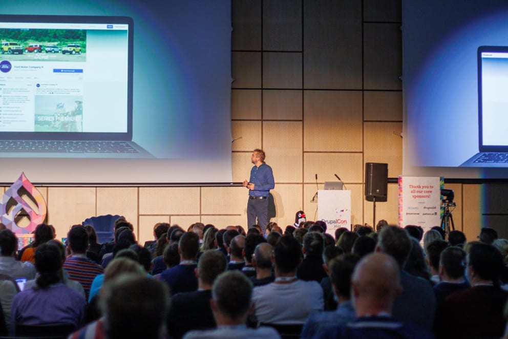 Man presenting into from an audience with a large screen behind him