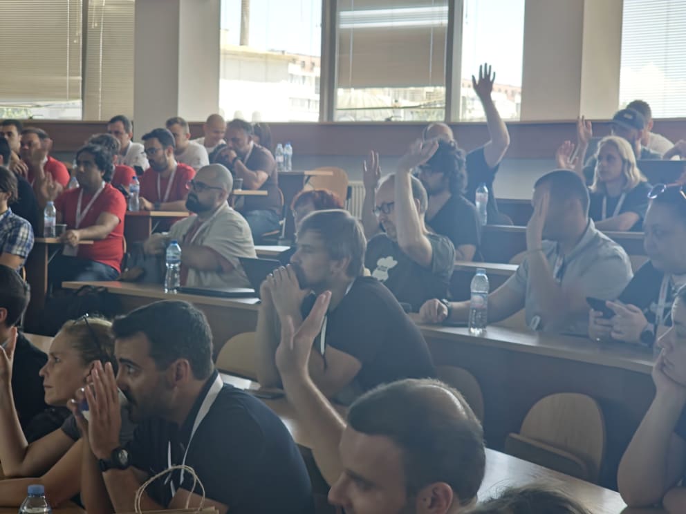 People sitting in a seminar, several with hands raised, appearing engaged and attentive.