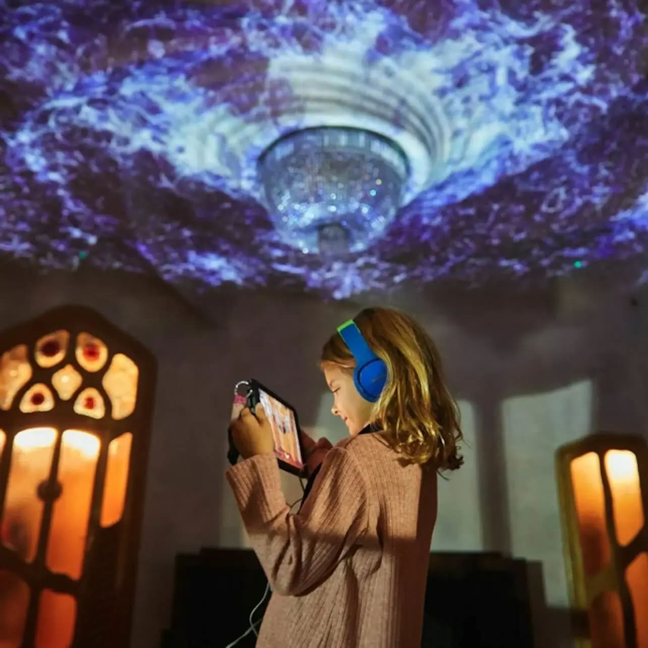 Un nen amb auriculars sosté una tauleta en una habitació amb decoració ornamentada i un sostre il·luminat per projeccions acolorides.