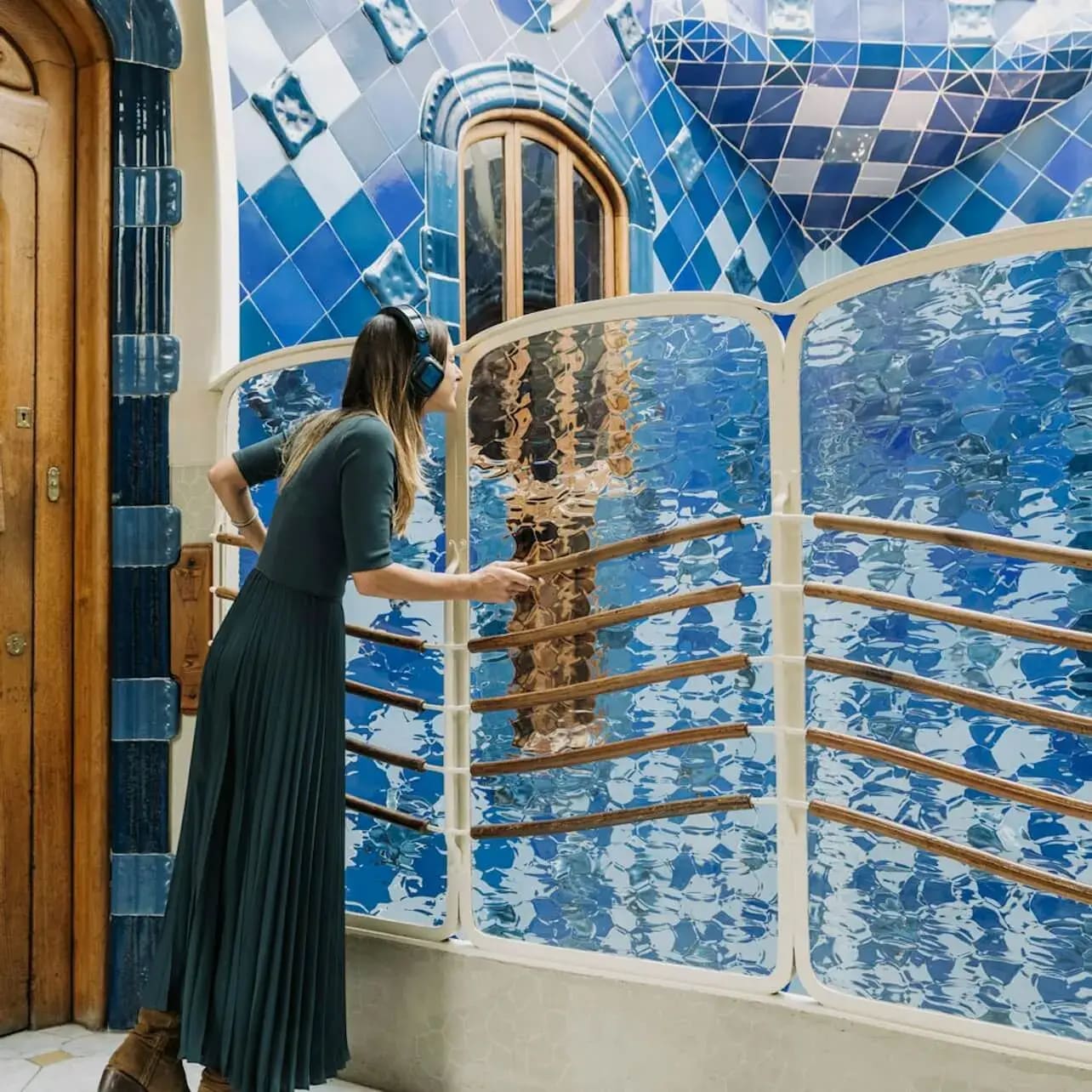 A woman in a long dress listens to headphones while looking over a wavy, reflective glass railing in a tiled, blue-toned space.