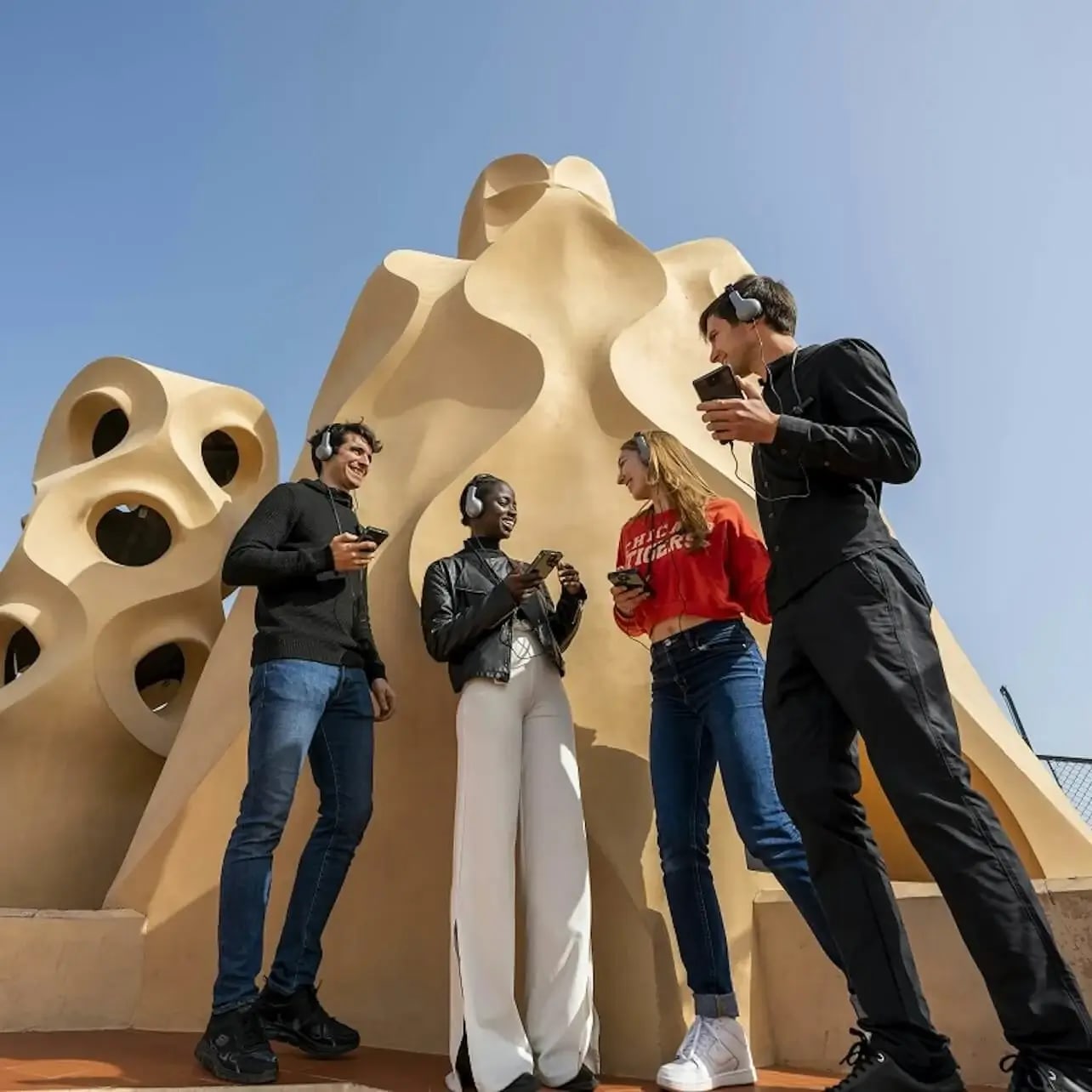 Quatre persones amb auriculars estan drets i conversen davant d'una estructura arquitectònica única amb formes orgàniques, sota un cel clar.