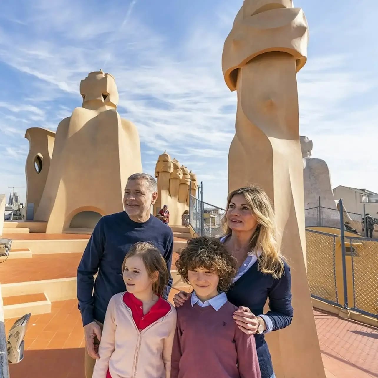 Una família de quatre persones està dreta en una terrassa amb estructures escultòriques en forma de xemeneia darrere seu sota un cel blau brillant.