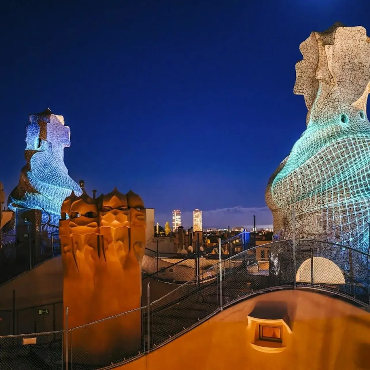 Vista del terrat de nit amb les ornamentades xemeneies i ventiladors de Gaudí il·luminats en diferents colors, sota un cel blau fosc clar.