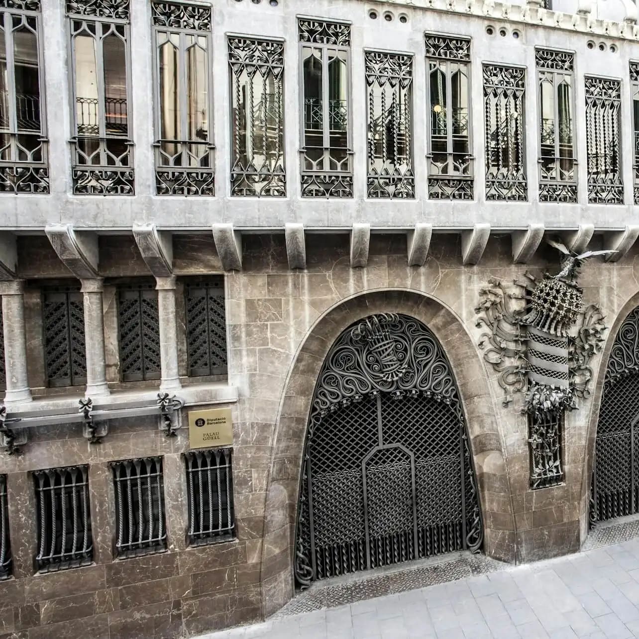Façana d'edifici ornamentada amb ferreria decorativa i pedra. Entrada arquejada i detalls intricats sobre les finestres. Un rètol diu "Gaudí".