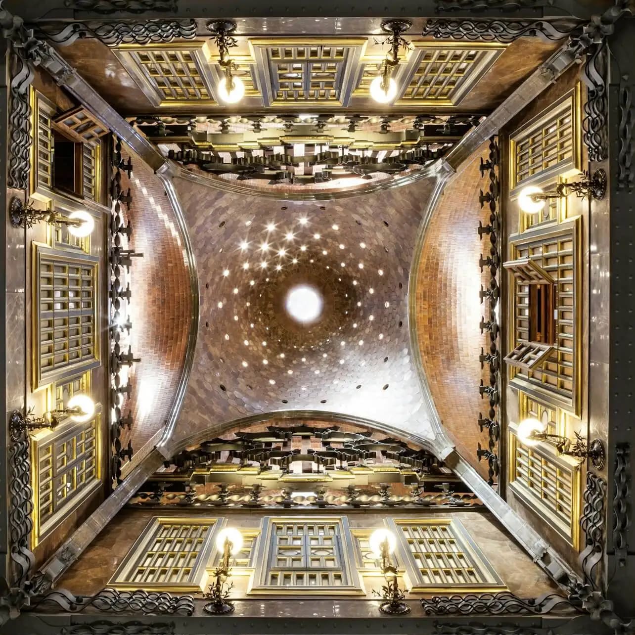 A symmetrical shot of an ornate, vaulted ceiling with decorative lights, windows, and intricate architectural detailing.