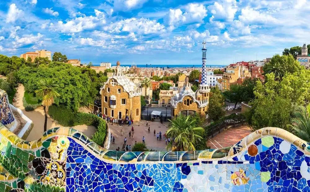 Vista panoràmica del Park Güell a Barcelona amb mosaics de colors en primer pla, dos edificis de formes fantàstiques i la ciutat al fons.