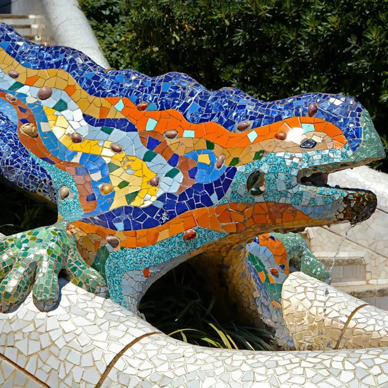 Escultura de lagarto de mosaico colorido con teselas verdes, azules y naranjas. Vegetación y escaleras al fondo.