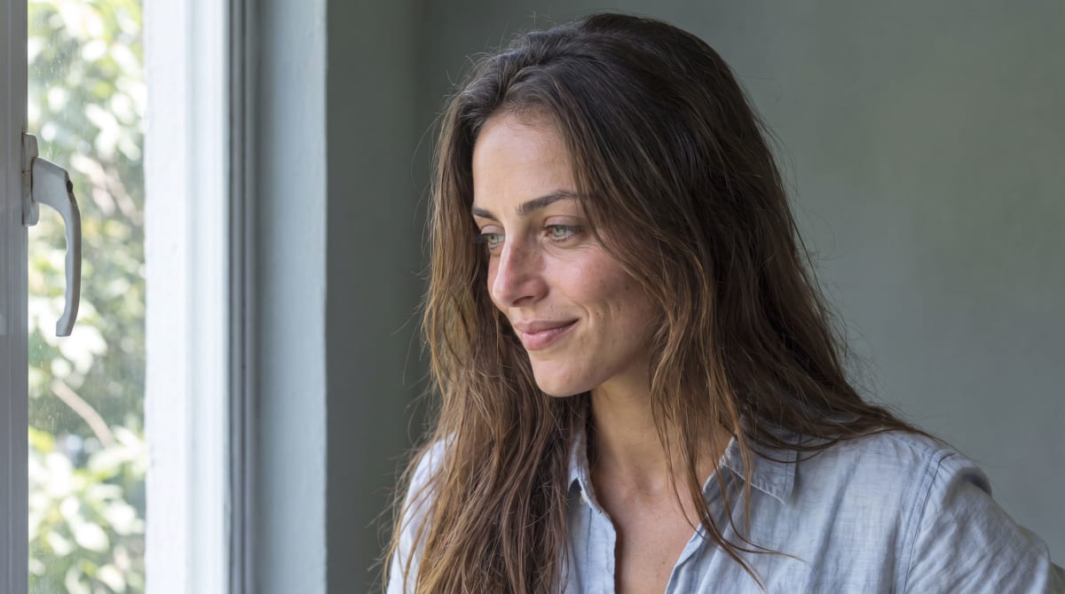 Close portrait of a fictional woman in her late twenties with long chestnut hair, light eyes and light olive skin, smiling slightly as she looks out of a window, in a pale blue shirt.