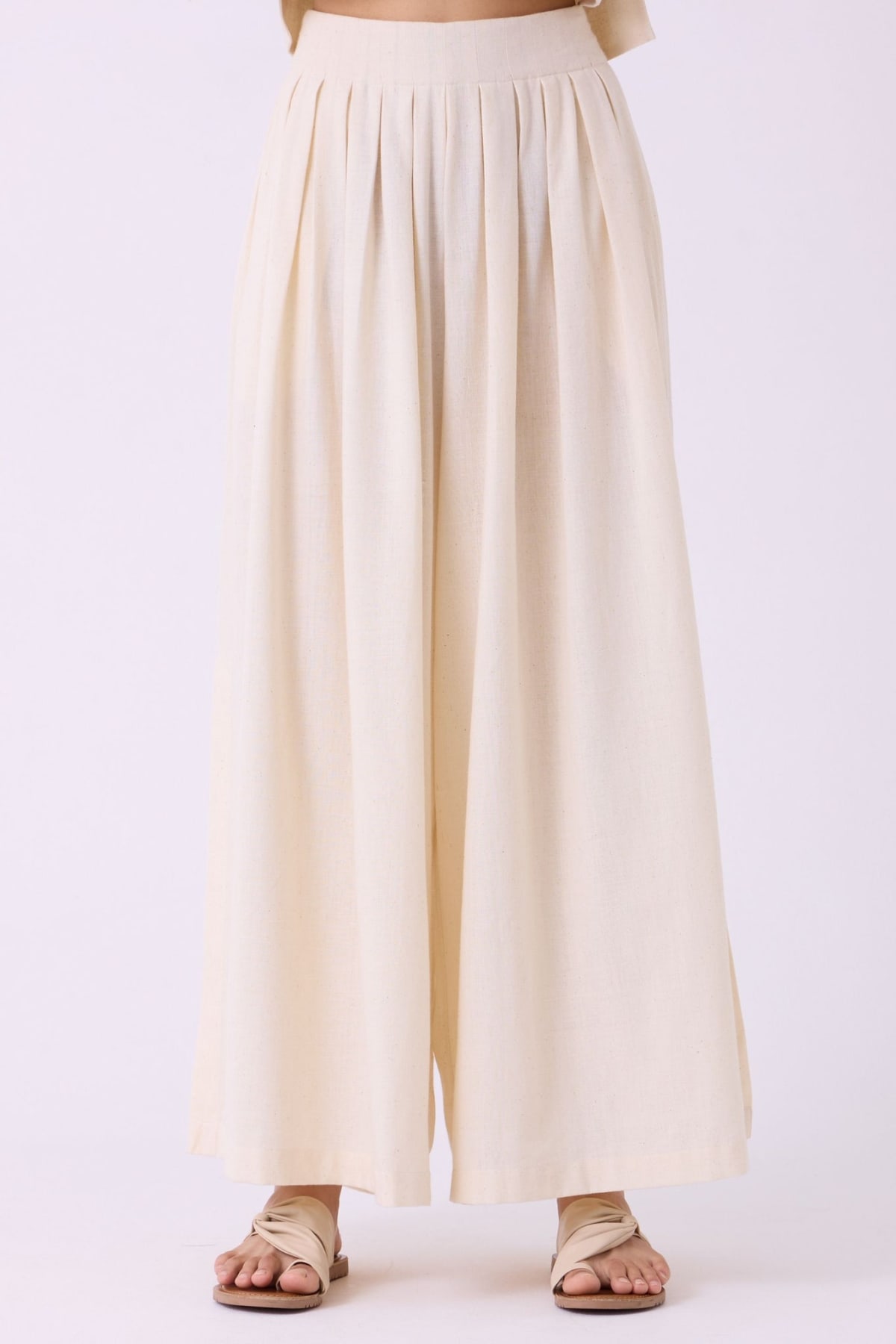 Official front product photograph of The Summer House Takumi trousers: off-white handwoven cotton with a broad waistband and narrow pleats falling into two wide legs, worn with sandals on a white studio background.
