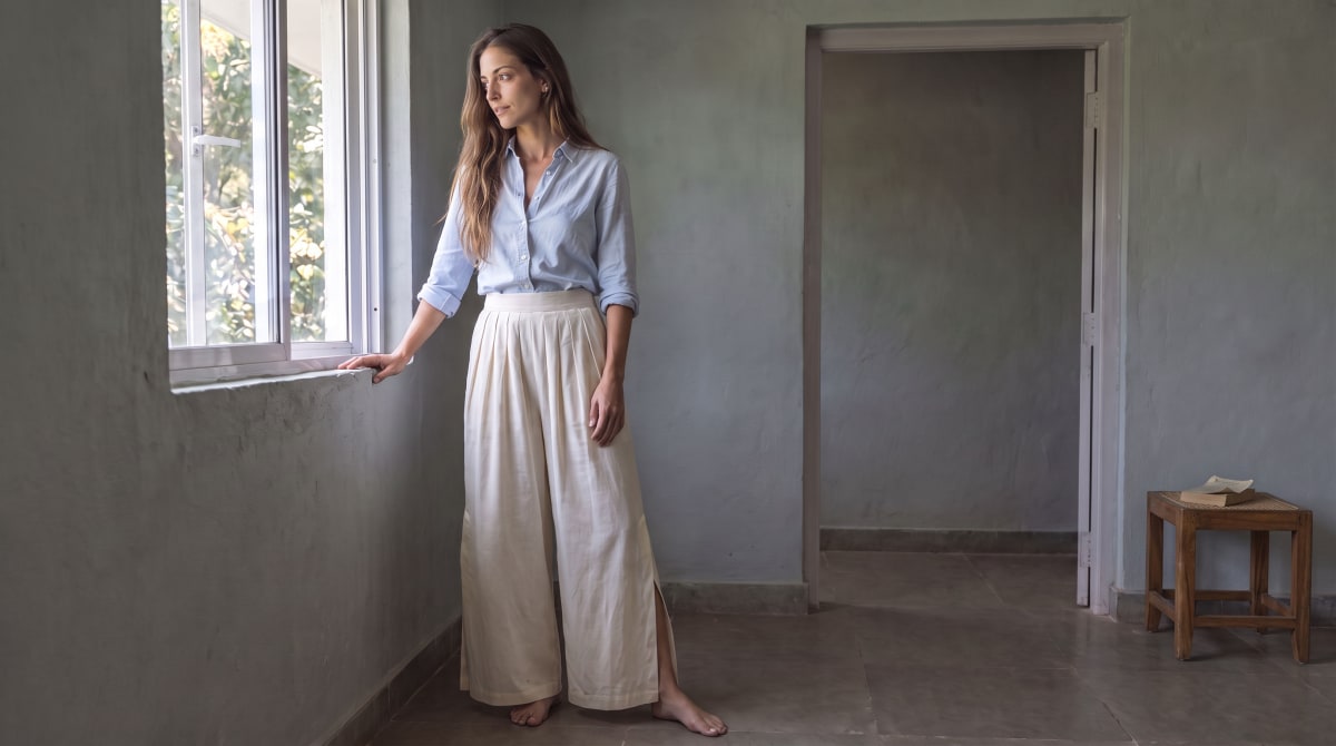 Room and light study: the woman in a pale blue shirt and off-white wide-leg trousers stands barefoot by an open window in a grey-green room, with a doorway and a wooden stool holding an open book to her right.