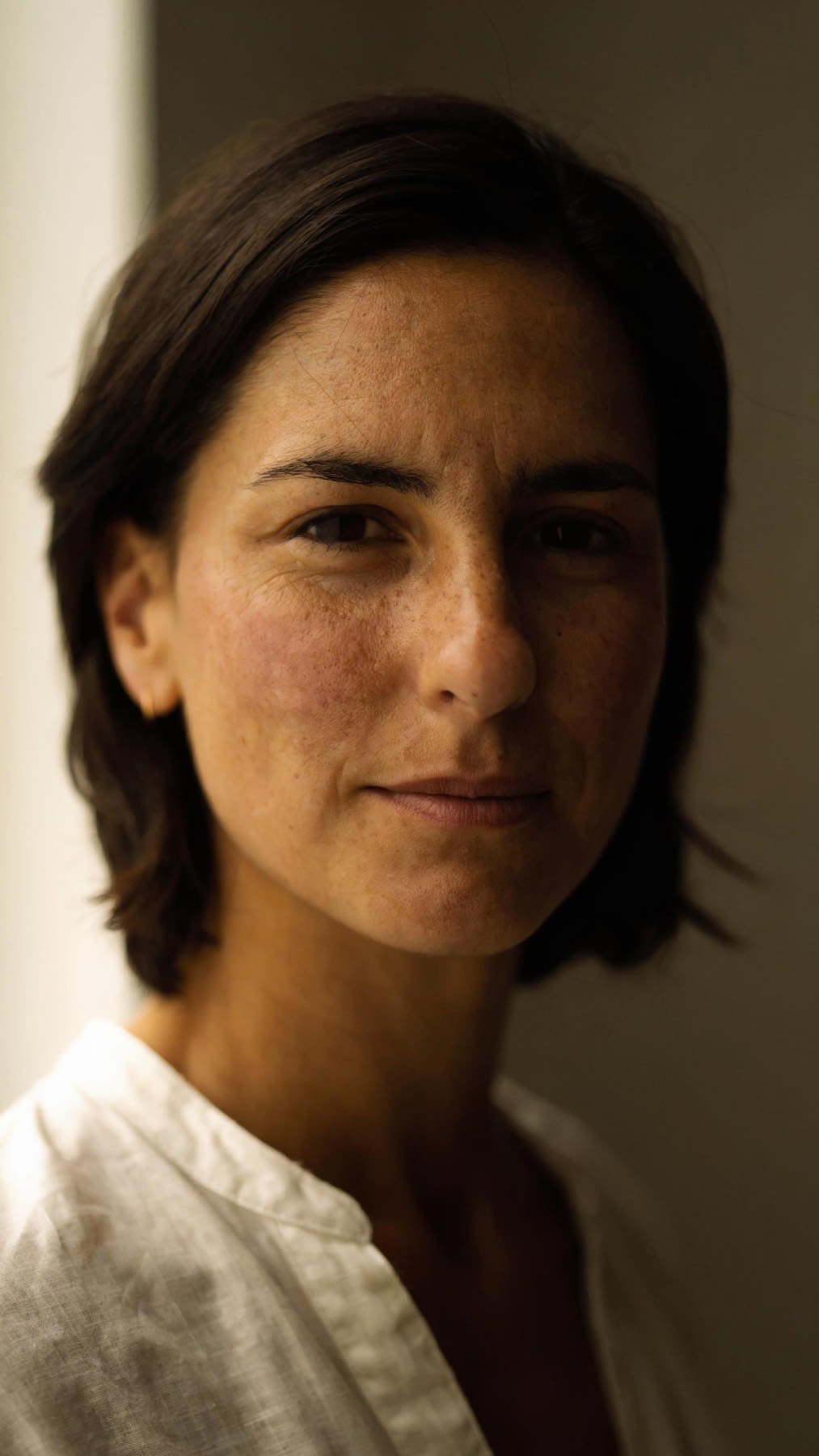Portrait of Lena, a fictional woman in her early thirties with dark shoulder-length hair tucked behind one ear, faint freckles and one small gold hoop, in an ivory linen shirt.