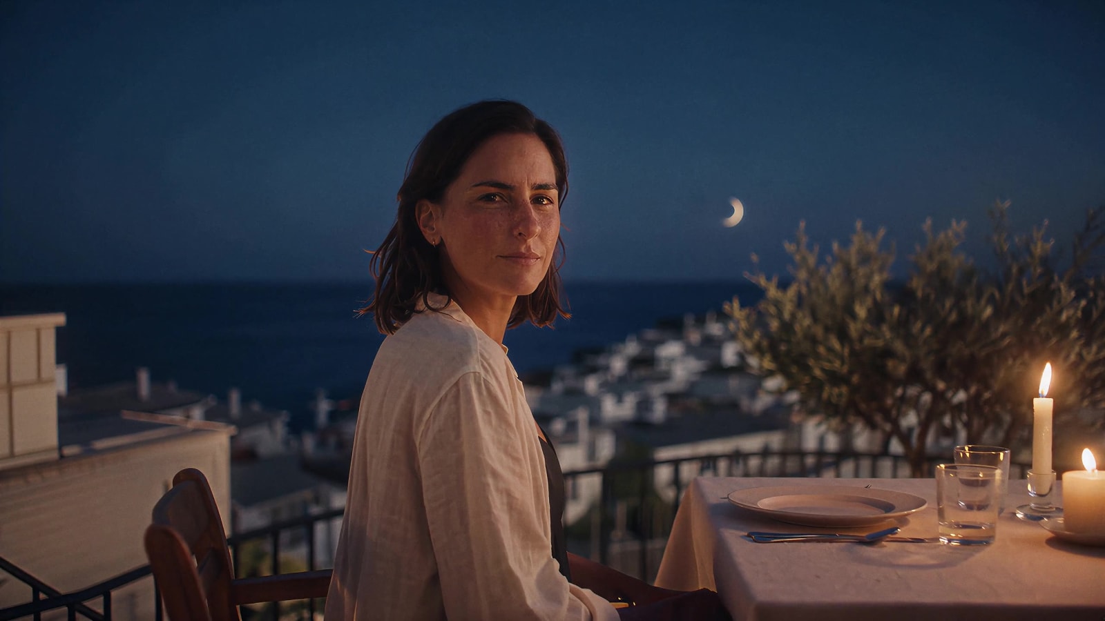 A table on a balcony at blue hour, above the lights of a village and a dark coastline.