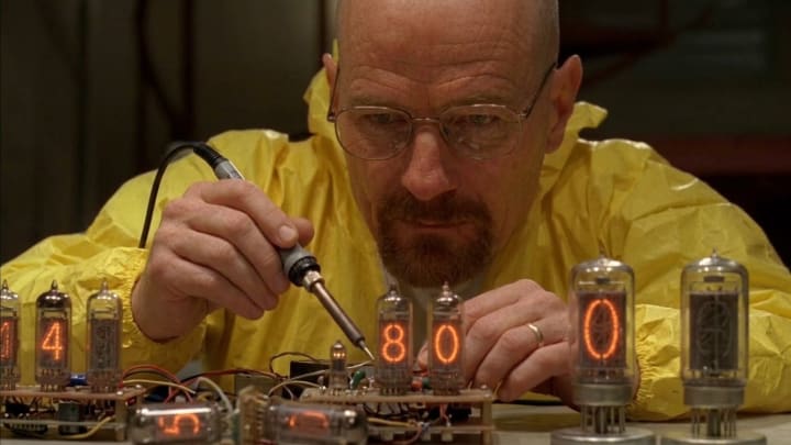 A man in a yellow protective suit soldering a Nixie tube clock with glowing digits