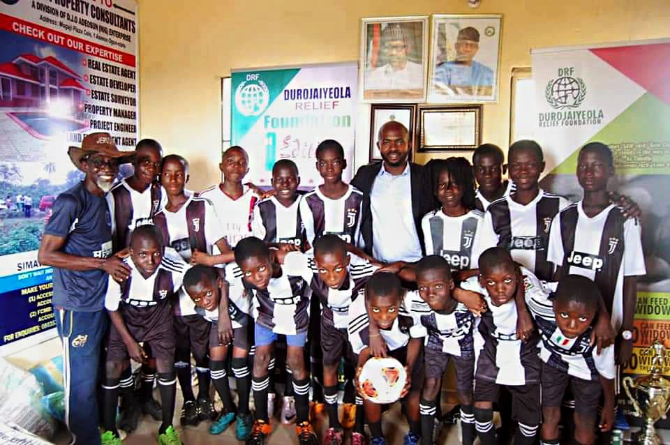 A youth football team in striped kits posing with coaches in front of Durojaiyeola Relief Foundation banners