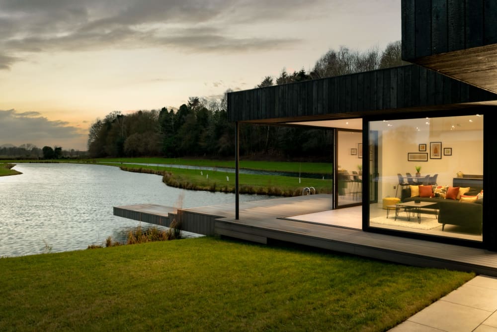 Modern lakeside home with floor-to-ceiling glass walls, dark timber cladding, wooden deck, and open-plan living space at dusk