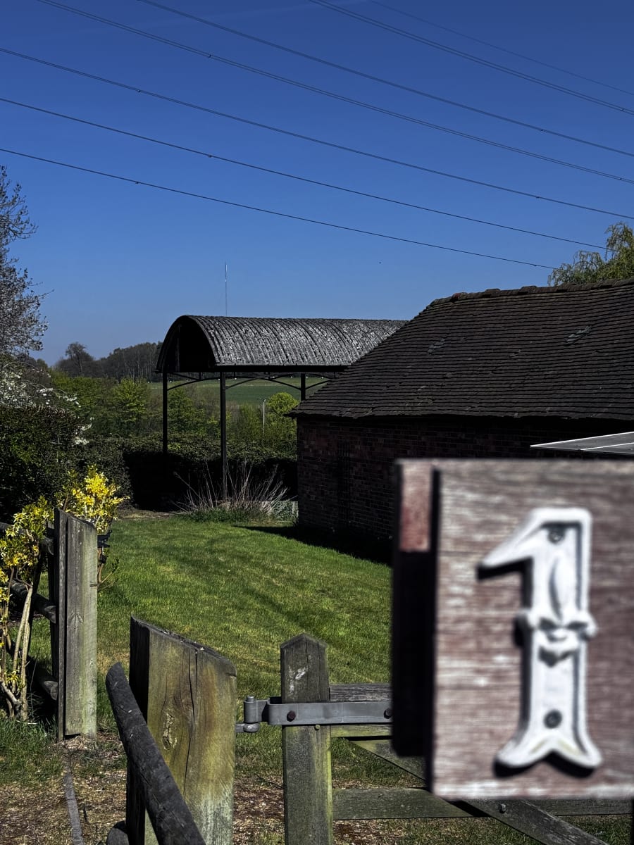 Rural property entrance with number 1 sign, curved roof barn structure, brick outbuilding, and green lawn under clear blue sky