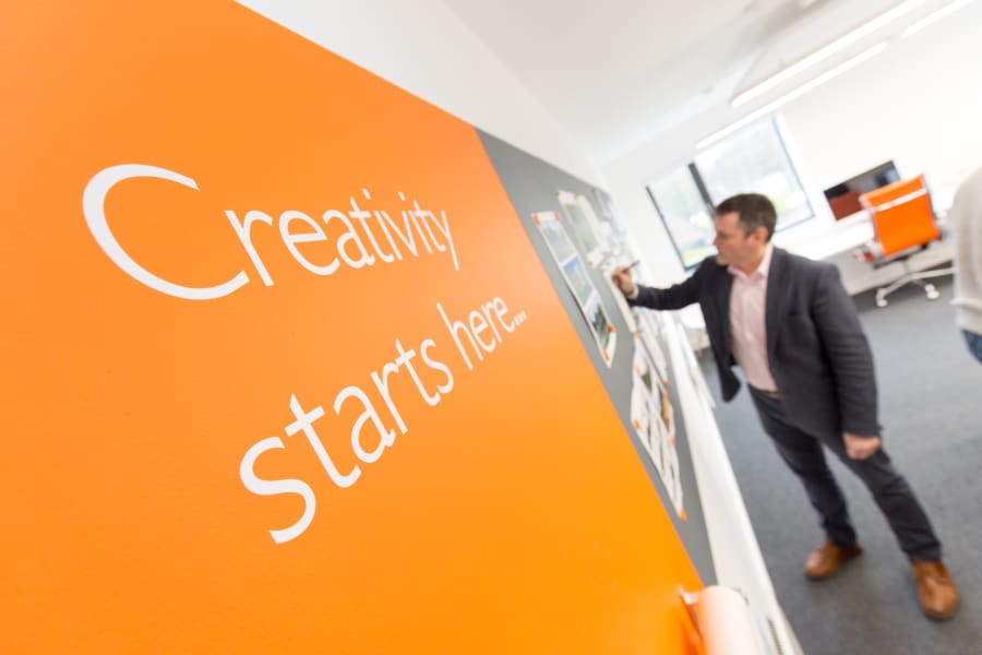 Man in a suit pins design sketches to a studio wall beside an orange "Creativity starts here" graphic
