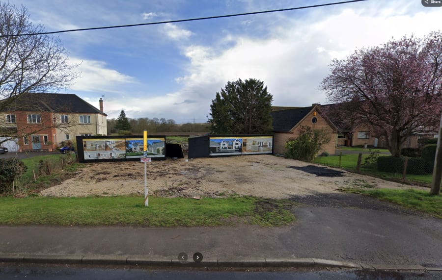 Vacant gravel building plot with development hoarding panels showing house renderings, flanked by existing homes on a residential street