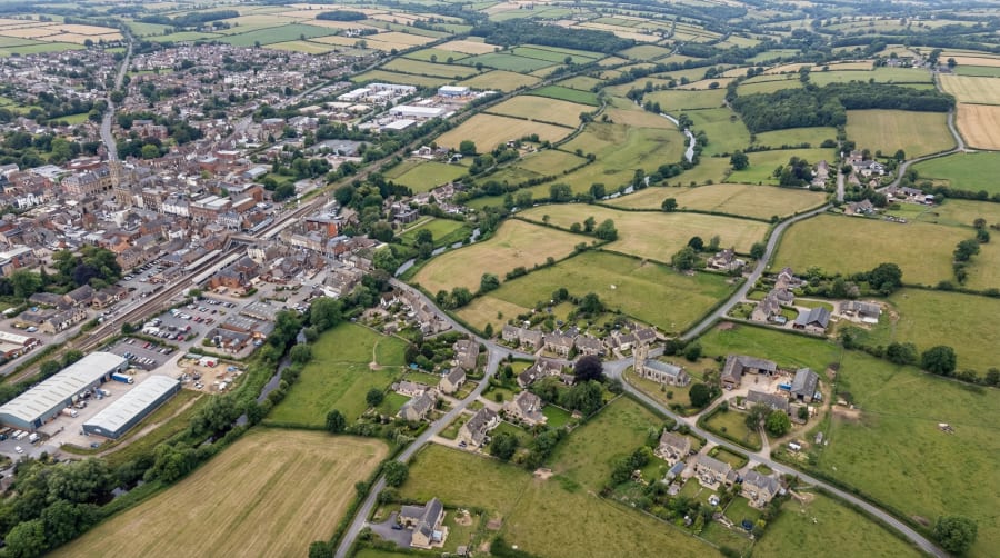 Aerial view of English market town with railway station, church spire, surrounding farmland and Cotswold stone villages