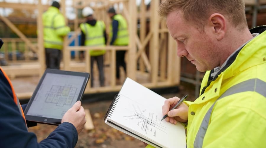 Site worker sketching a construction detail while a colleague holds a tablet with floor plans, timber frame building behind