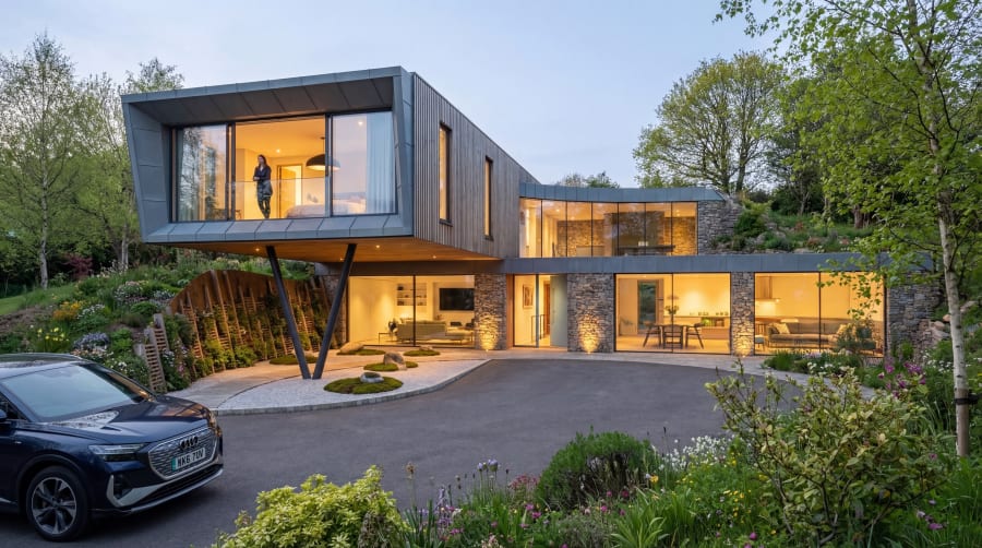 Cantilevered dark-timber upper floor projects over a stone-clad lower level at dusk, illuminated from within
