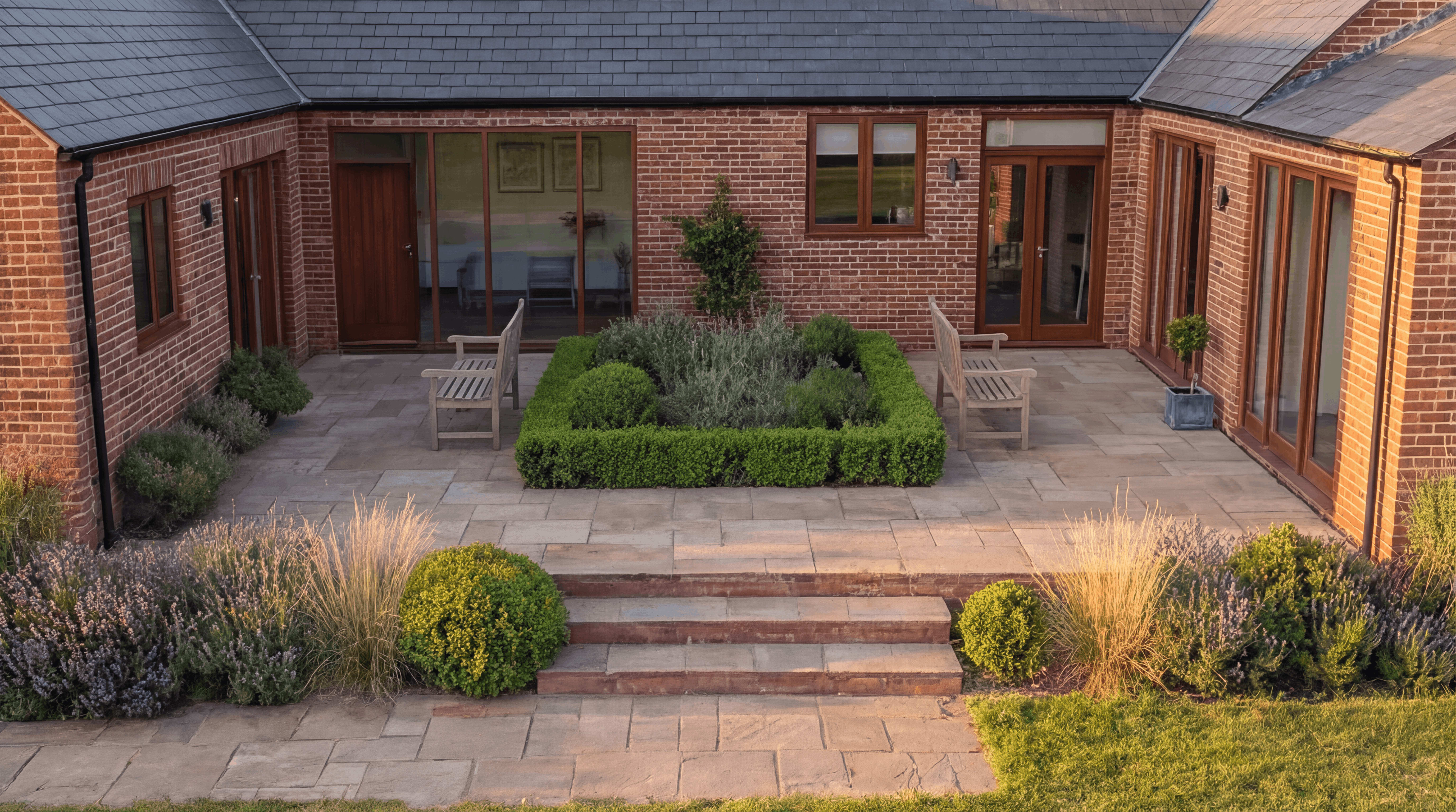 Brick courtyard of a converted barn-style home with slate roof, timber-framed doors, and a boxwood-hedged garden bed flanked by teak benches