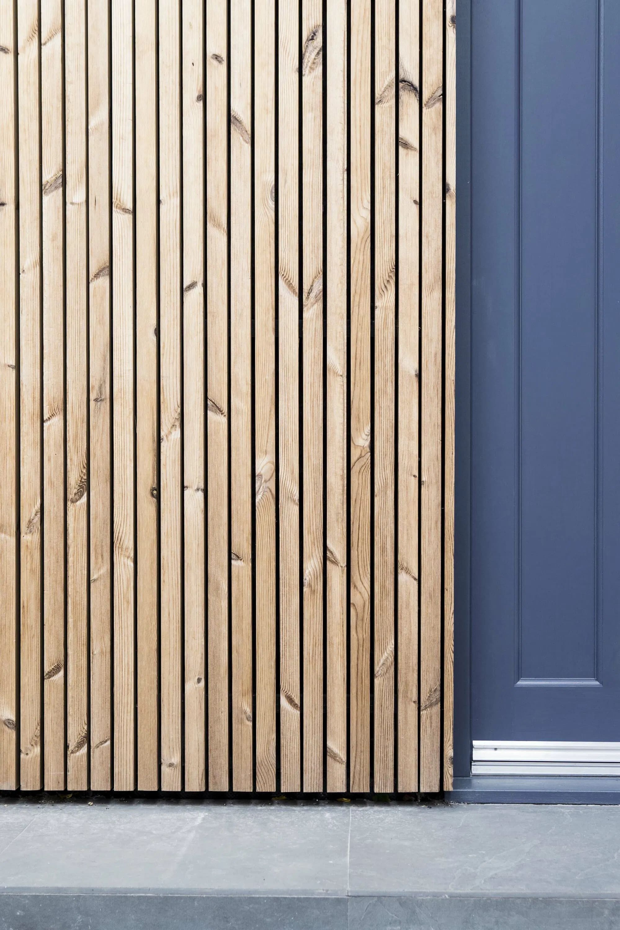 Vertical natural wood slat cladding detail next to dark blue-grey painted door frame with stone paving below
