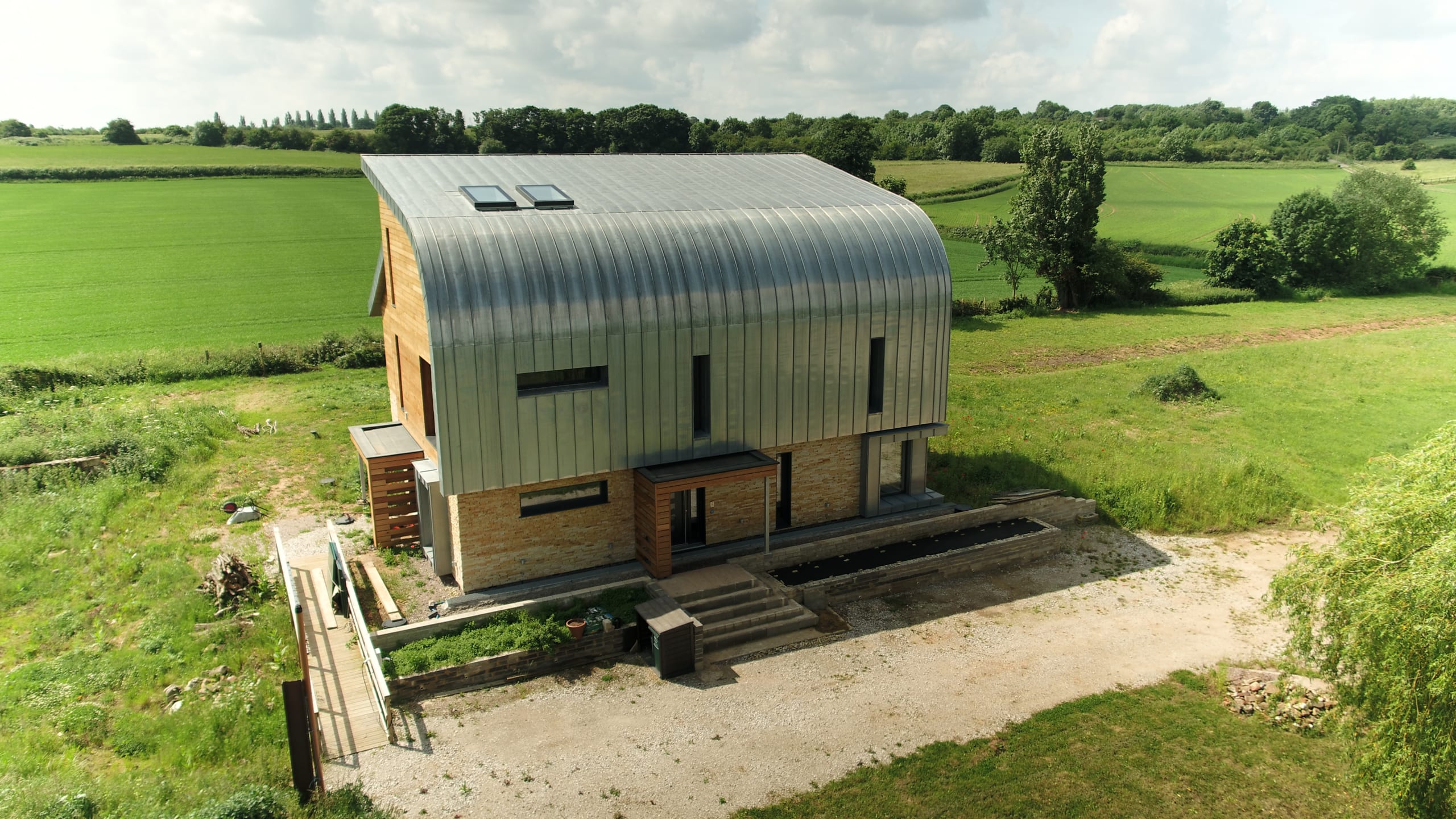 Modern rural home with curved zinc roof, timber cladding, and stone base set in green countryside landscape